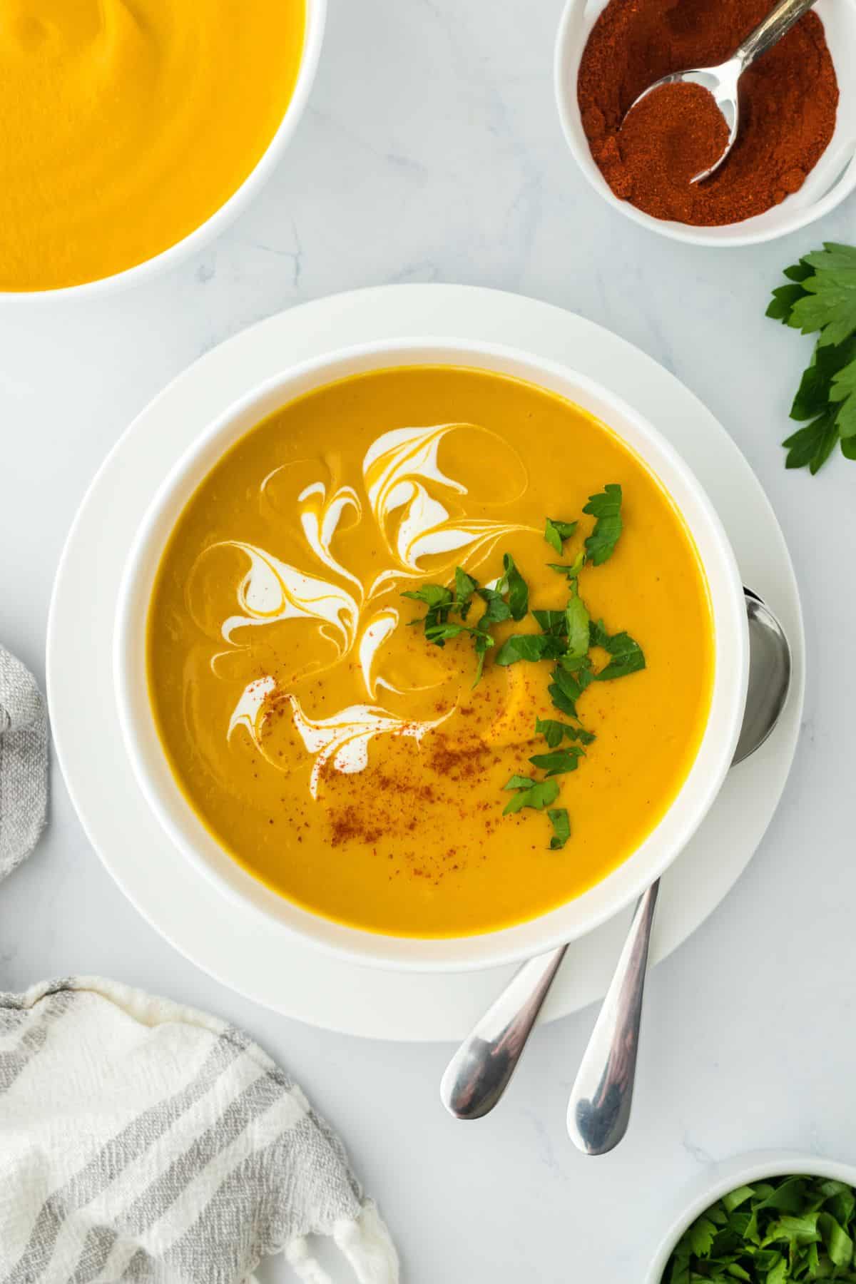 A bowl of sweet potato soup with cream swirls, fresh herbs, and a dusting of spices, surrounded by bowls of garnish, more soup, and a cloth napkin