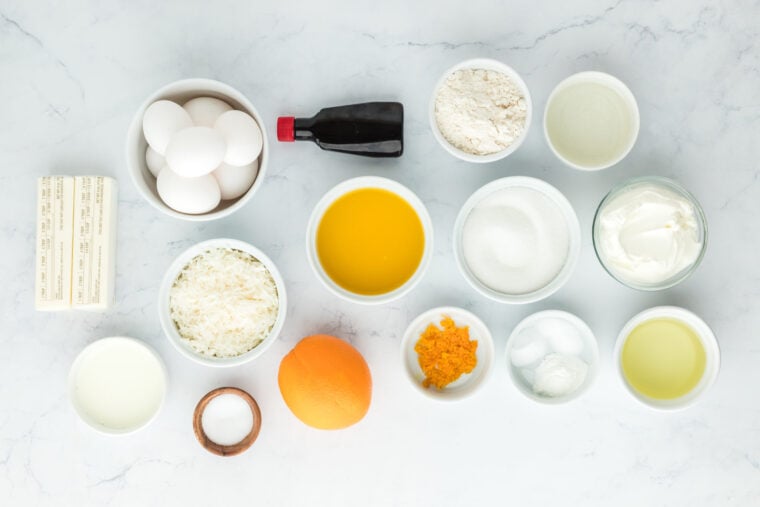 Coconut, eggs, orange juice, vanilla, and other ingredients in white bowls on white countertop