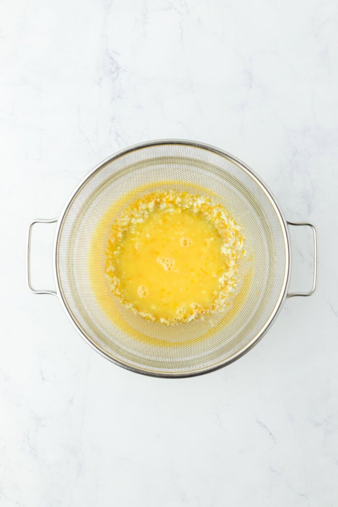 Orange curd being pushed through a fine mesh strainer on white countertop
