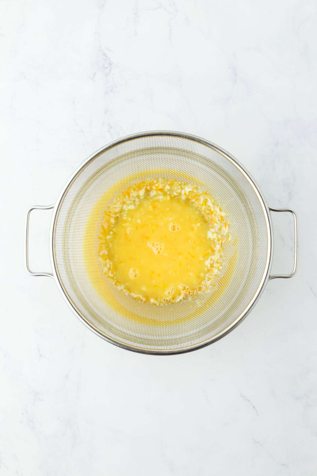 Orange curd being pushed through a fine mesh strainer on white countertop