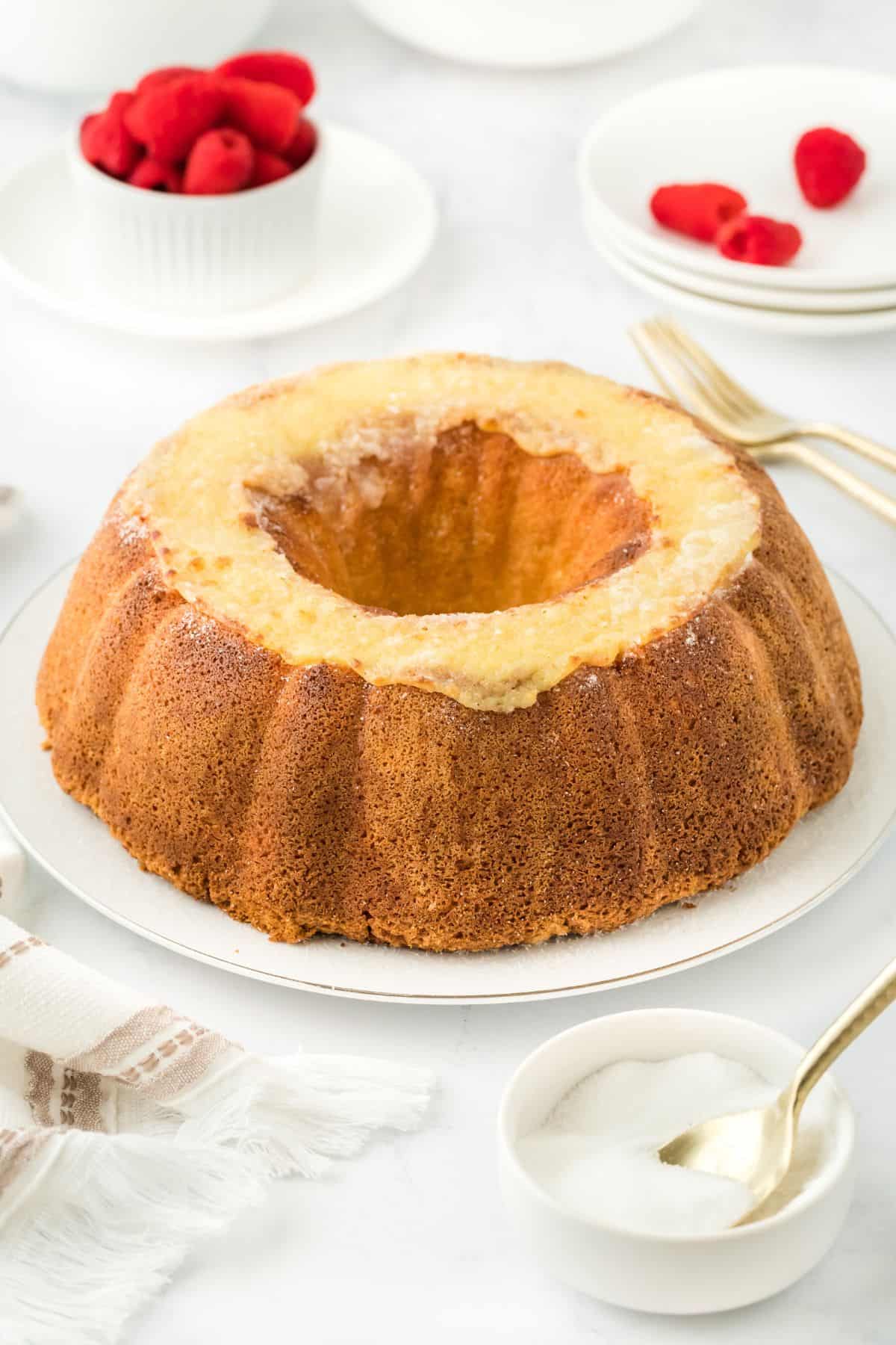 A whole creme brulee pound cake displayed on a white plate, surrounded by tableware and raspberries and a small bowl of sugar with a spoon in it