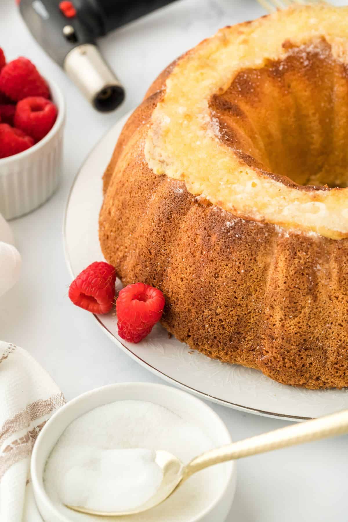 Angled view of the whole creme brulee pound cake, accompanied by gold utensils, a bowl of sugar, a blowtorch, and fresh raspberries