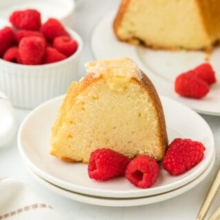 Close up of a slice of creme brulee pound cake with a golden crust, garnished with fresh raspberries on a white plate, with the rest of the cake and more raspberries in the background