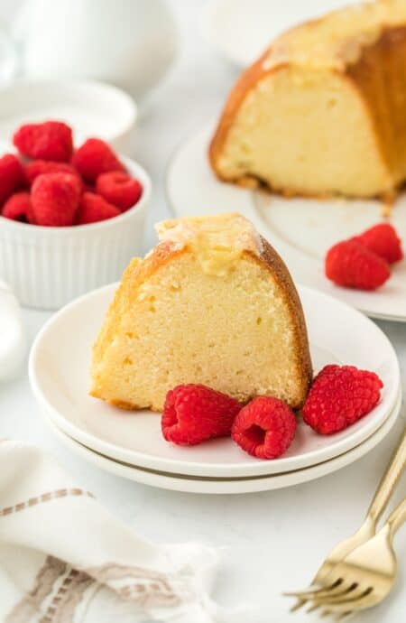 Close up of a slice of creme brulee pound cake with a golden crust, garnished with fresh raspberries on a white plate, with the rest of the cake and more raspberries in the background