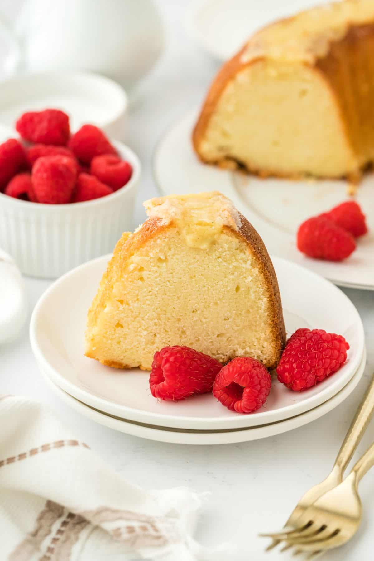 Close up of a slice of creme brulee pound cake with a golden crust, garnished with fresh raspberries on a white plate, with the rest of the cake and more raspberries in the background