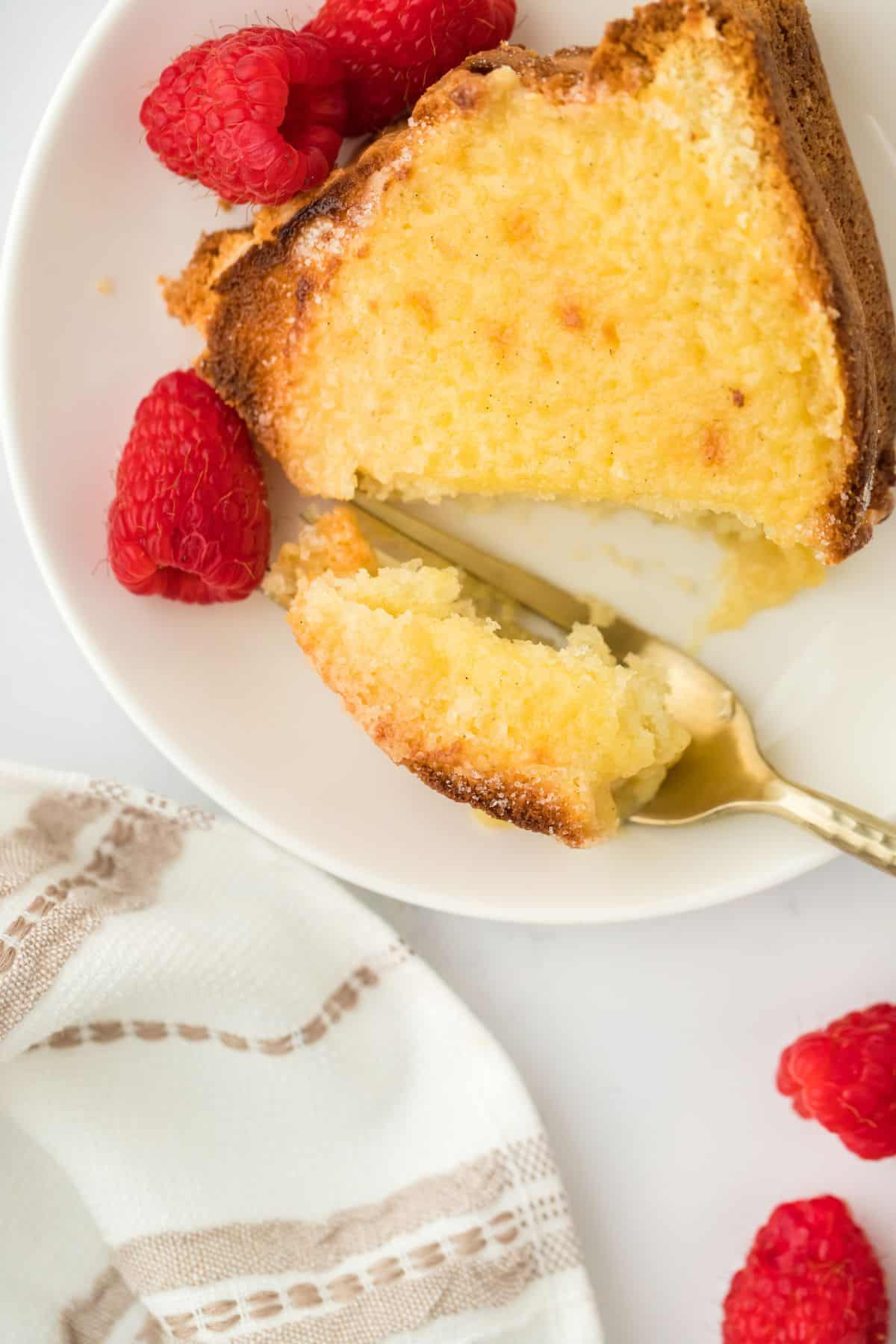 A single slice of creme brulee pound cake with a bite taken out, placed on a plate with a gold fork, surrounded by raspberries and a napkin