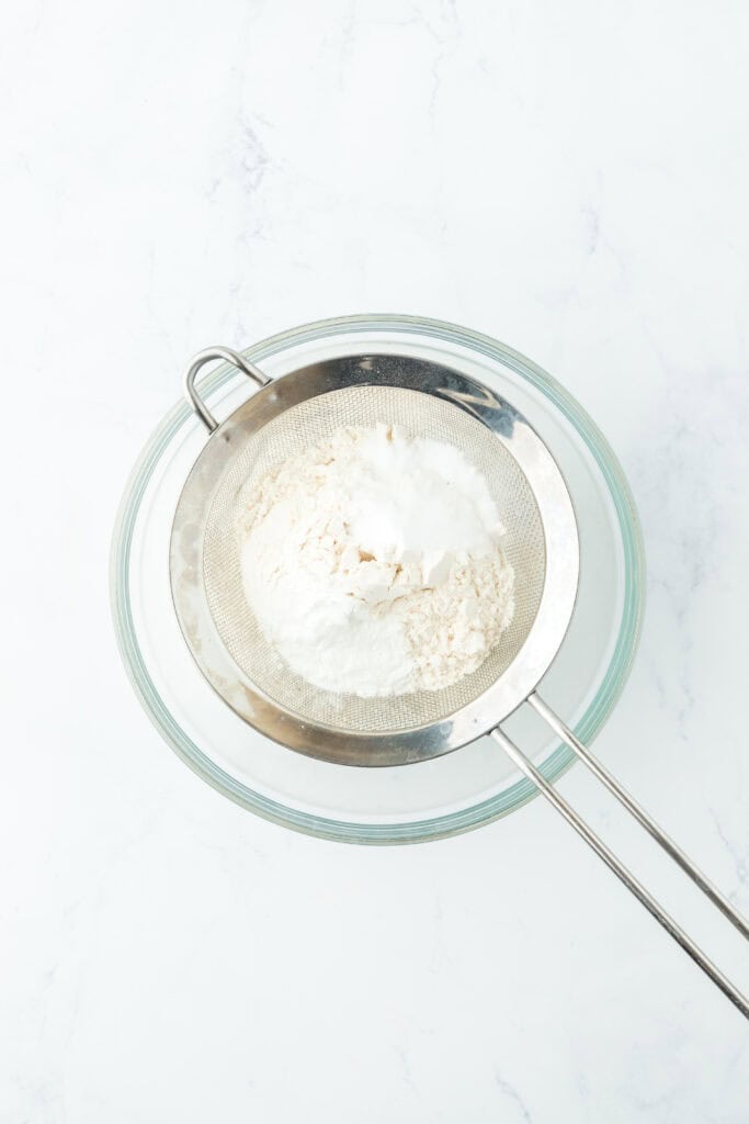Flour, baking soda, baking powder and salt being sifted into a bowl on white countertop