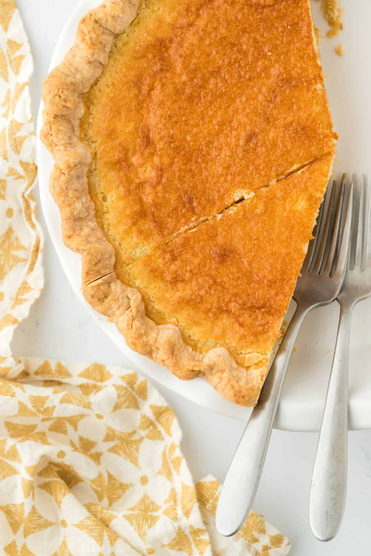 Overhead shot of a golden-brown double crusted sweet potato pie, partially sliced, and placed on a white plate with a yellow patterned cloth and two forks next to it