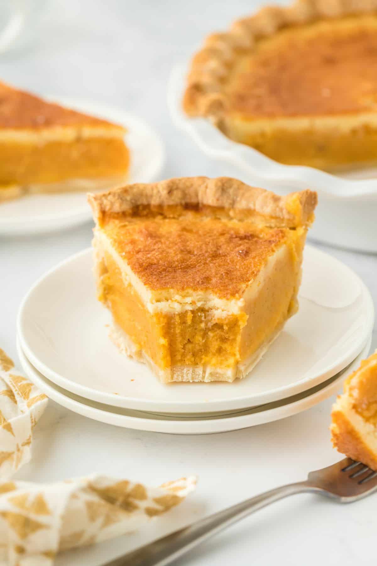 A slice of double crusted sweet potato on a small white plate with a bite taken off, showing layers of smooth texture, with the whole pie in the background