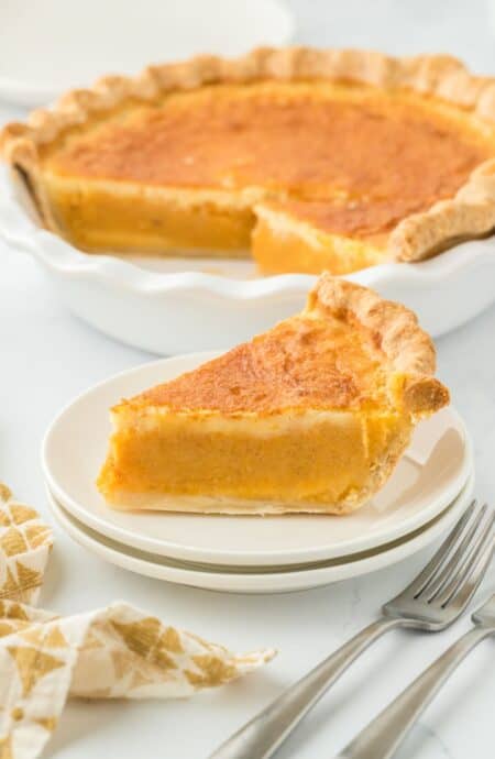 A slice of double crusted sweet potato pie on a small white plate, with the whole pie in the background