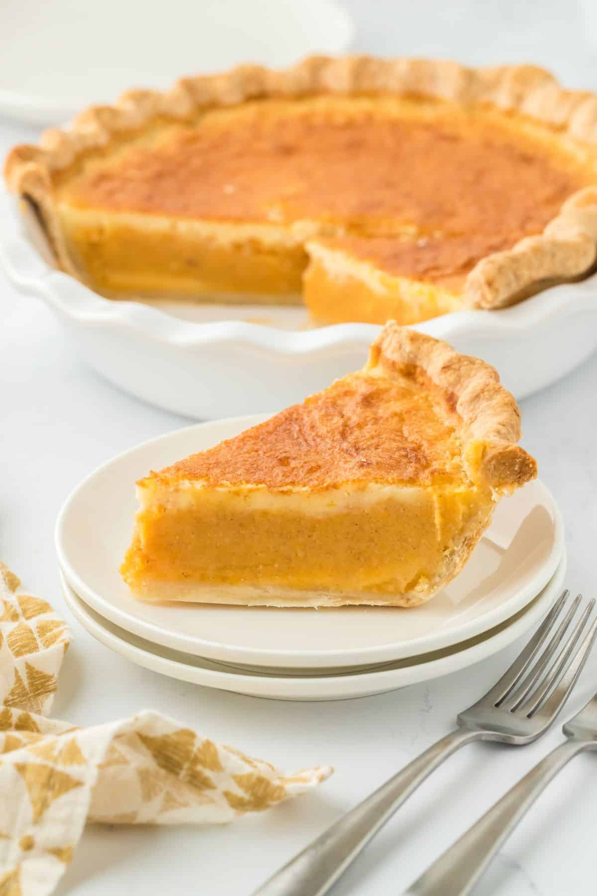 A slice of double crusted sweet potato pie on a small white plate, with the whole pie in the background