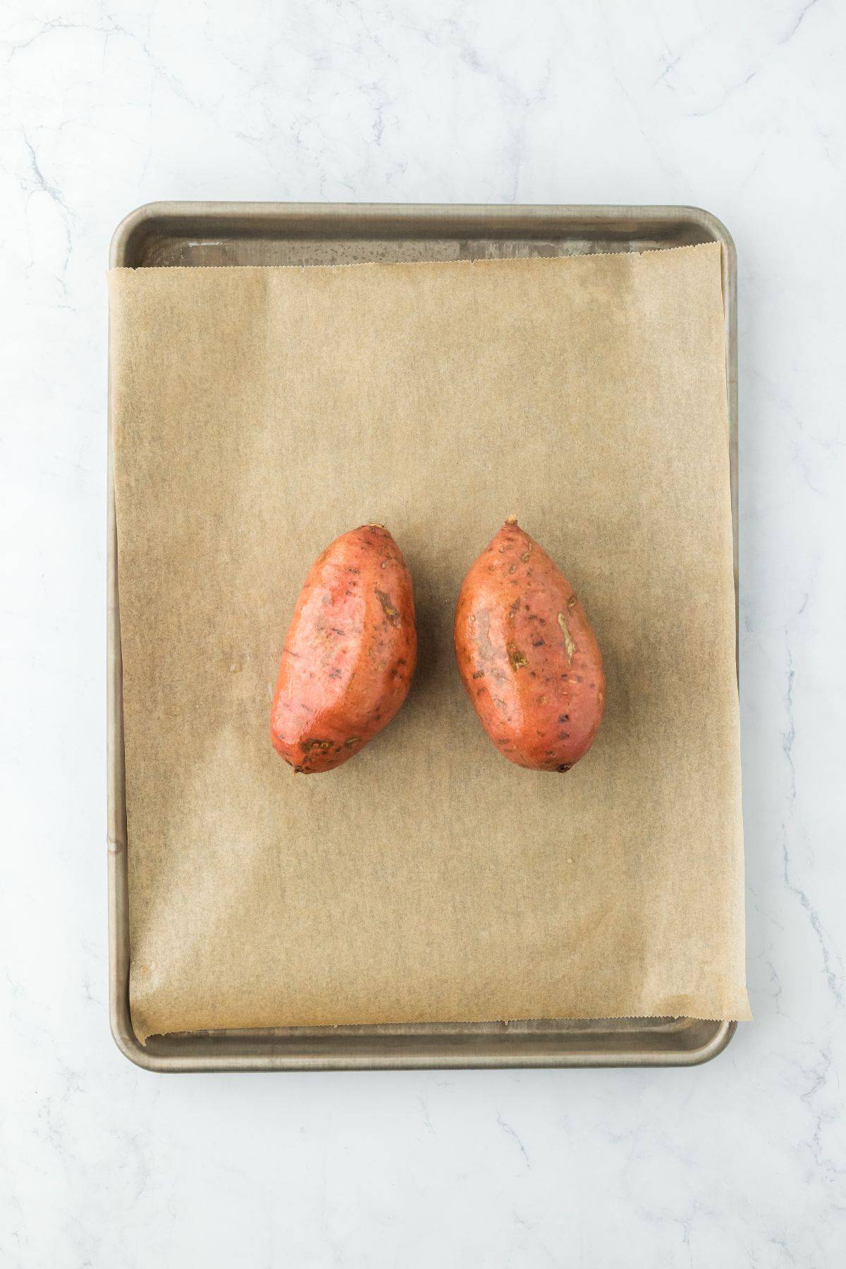 A tray with two whole sweet potatoes placed on parchment paper, ready for roasting