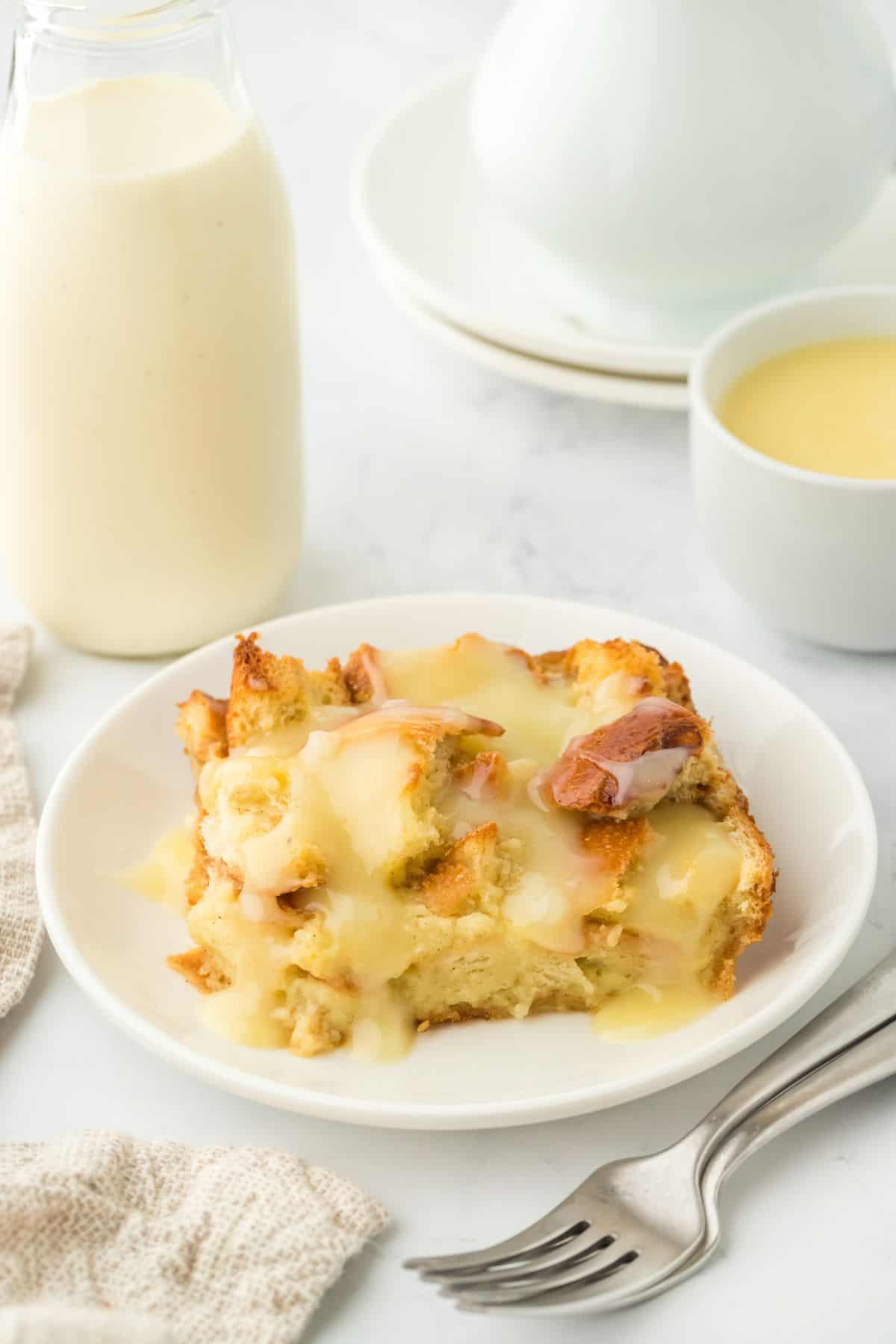 A serving of eggnog bread pudding on a white plate with vanilla sauce, accompanied by a bottle of eggnog and a cup of sauce in the background