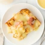 Overhead shot of a serving of eggnog bread pudding on a white plate, surrounded by a fork, a napkin, and a cup of sauce nearby