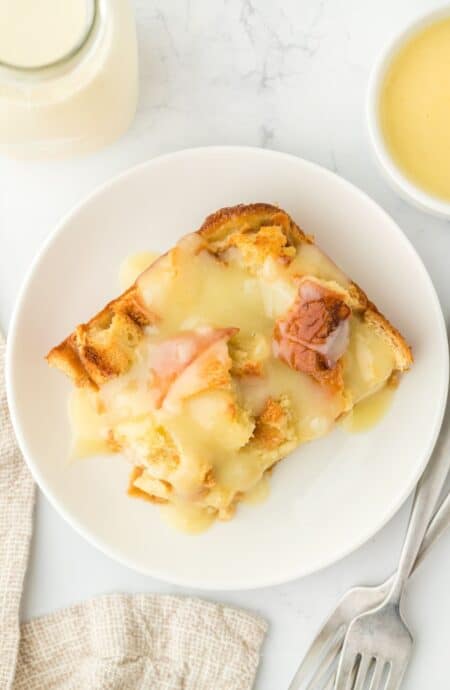 Overhead shot of a serving of eggnog bread pudding on a white plate, surrounded by a fork, a napkin, and a cup of sauce nearby