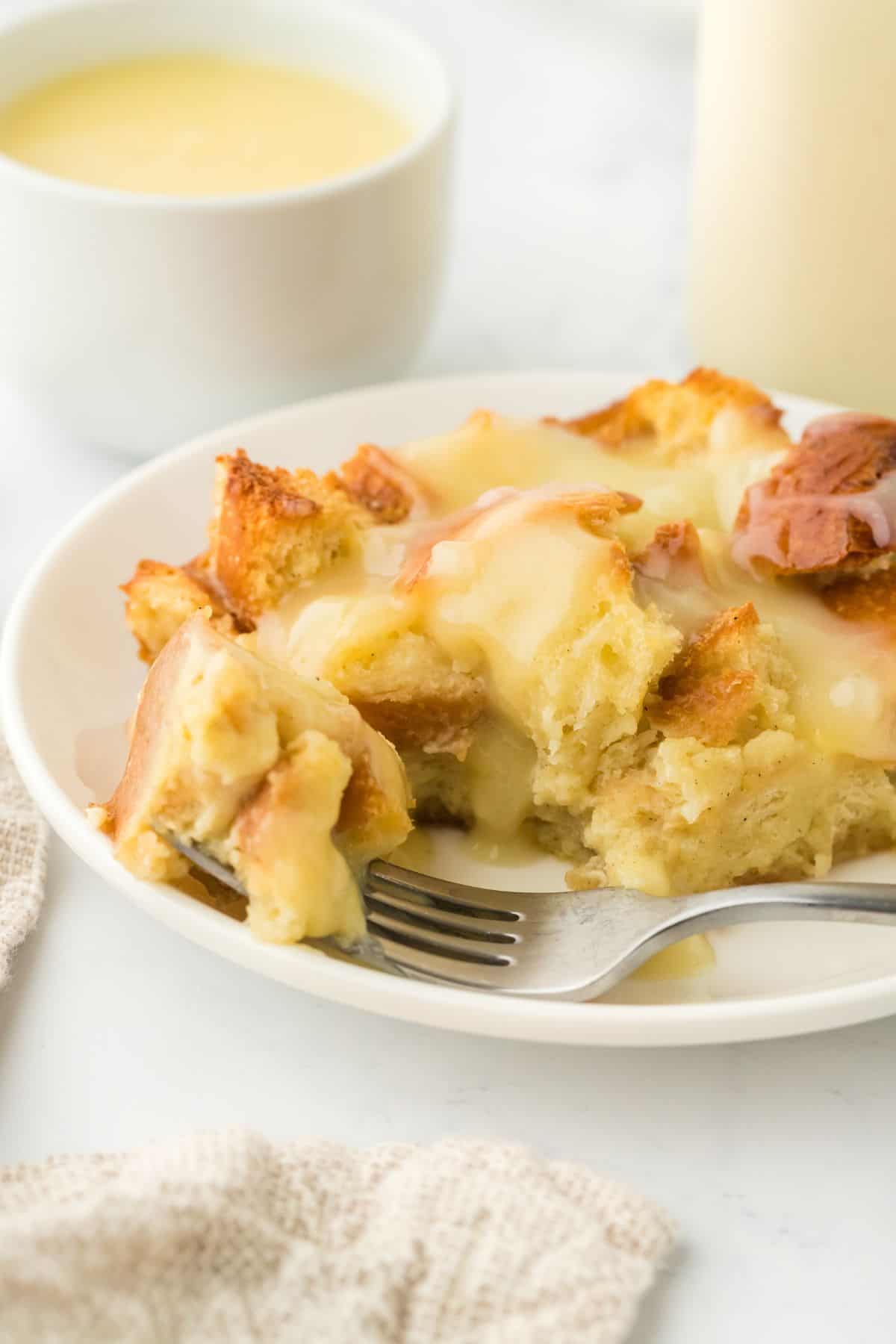 Closeup of eggnog bread pudding with a fork taking a bite and a bowl with vanilla sauce in the background