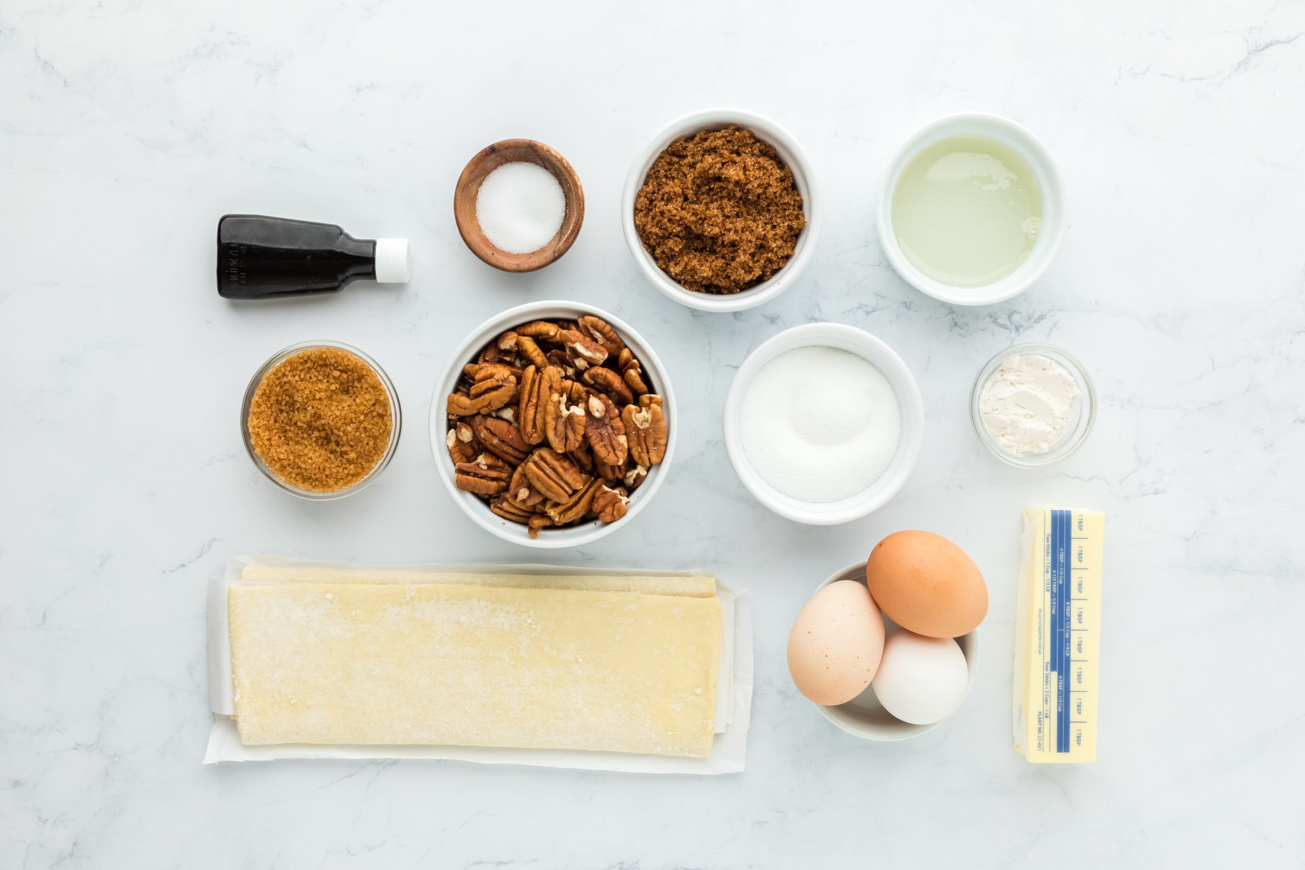 Puff pastry, pecans, sugars, vanilla, eggs, butter, salt, corn syrup and flour on a white countertop