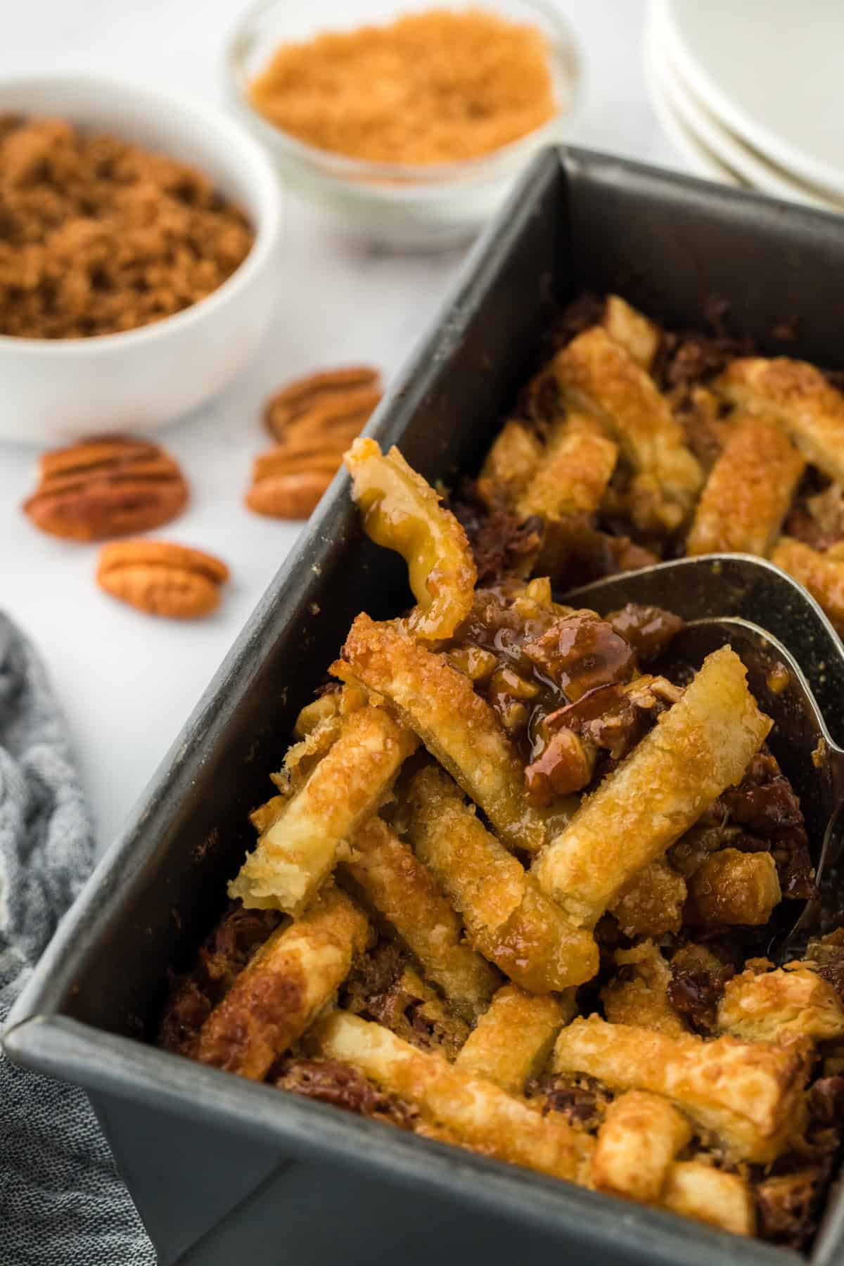 A close up of a spoon digging into pecan pie cobbler recipe on white countertop with pecans and brown sugar in background