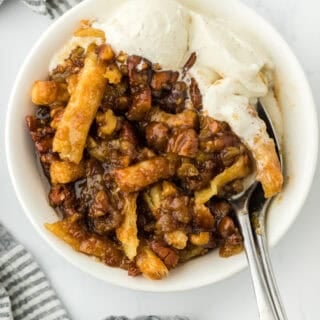 A white bowl of pecan cobbler with vanilla ice cream and spoon digging in on white countertop with striped napkin