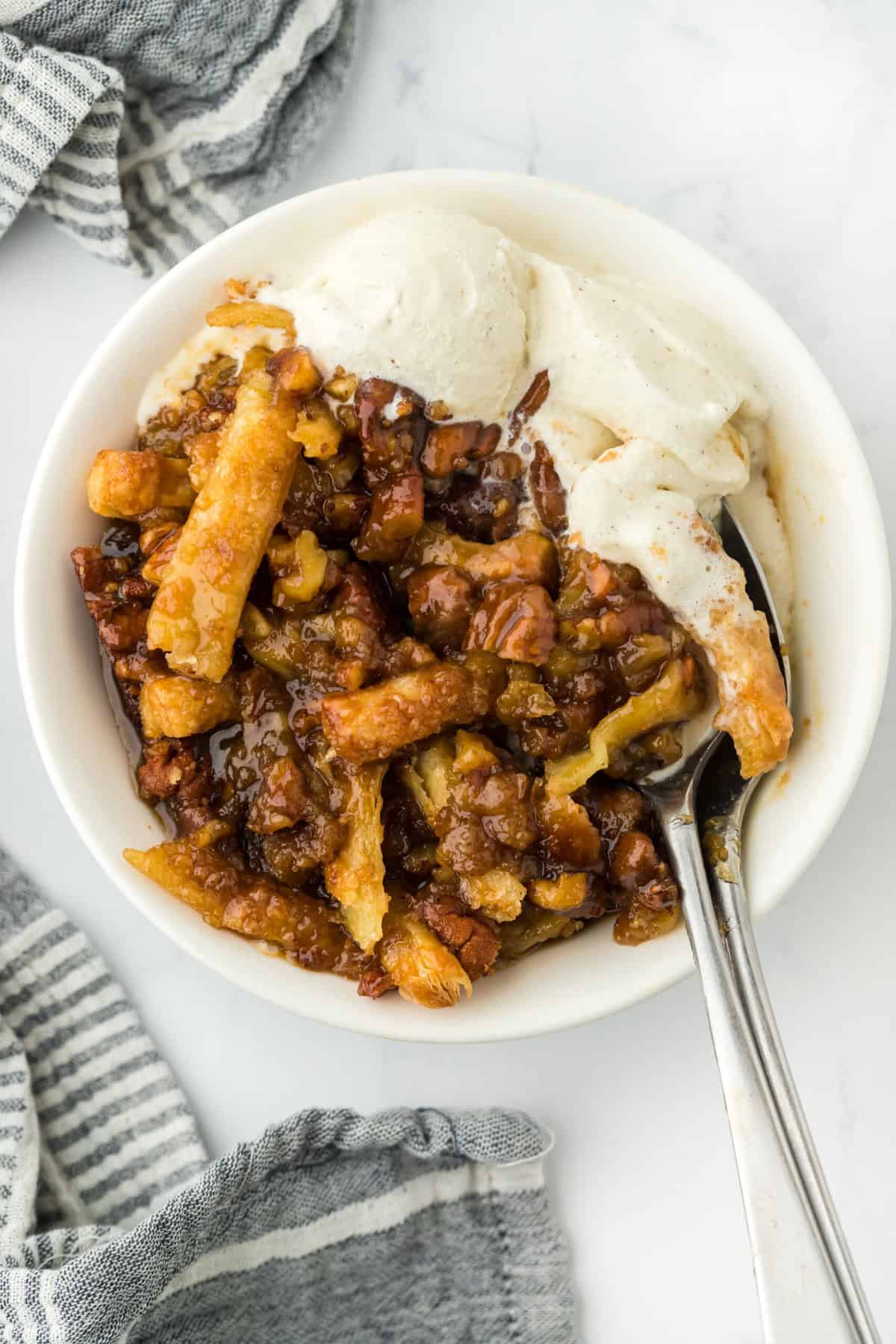 A white bowl of pecan cobbler with vanilla ice cream and spoon digging in on white countertop with striped napkin