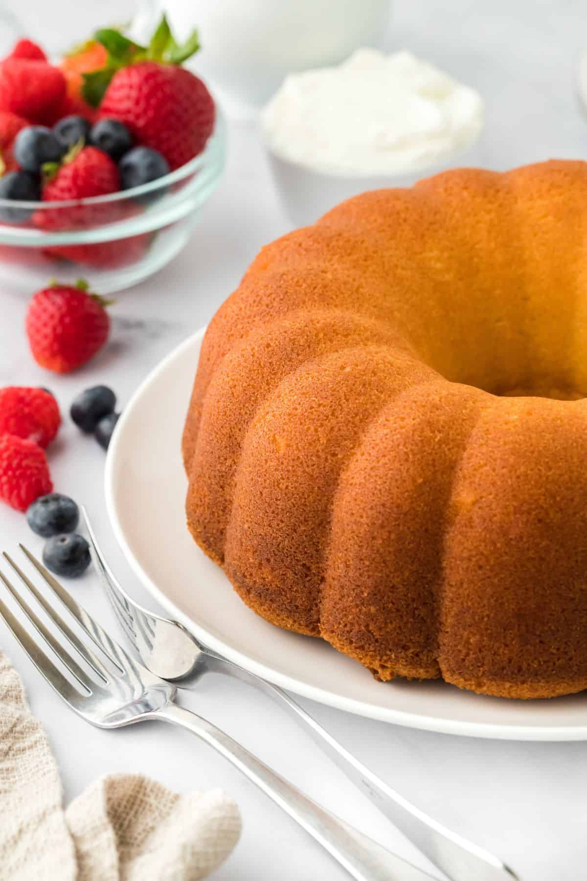 A whole whipping cream bundt cake displayed on a white plate, surrounded by fresh strawberries, blueberries, and a bowl of whipped cream in the background