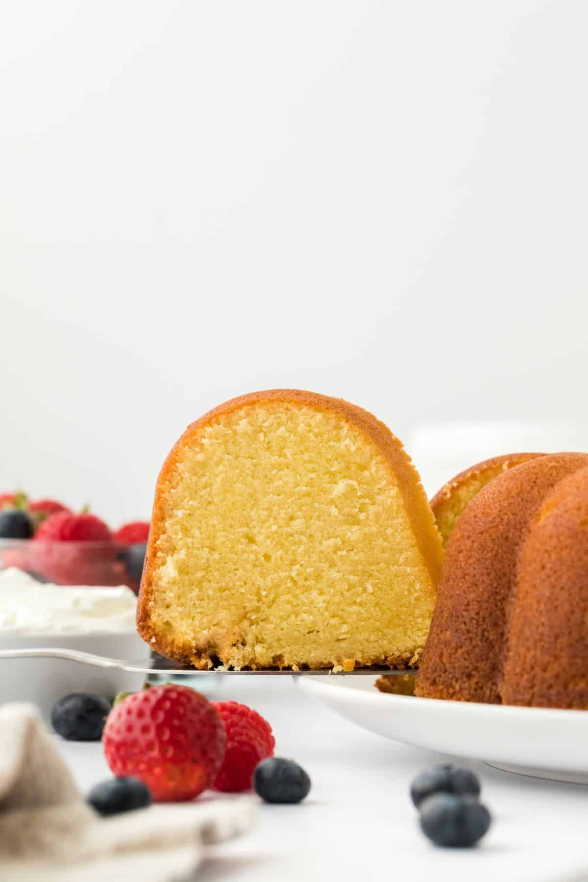 A close up of a slice of whipping cream pound cake being lifted with a cake server, with a background of fresh berries