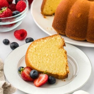 A slice of whipping cream pound cake served on a plate, garnished with fresh strawberries and blueberries, with the rest of the cake and a bowl with more fruit in the background