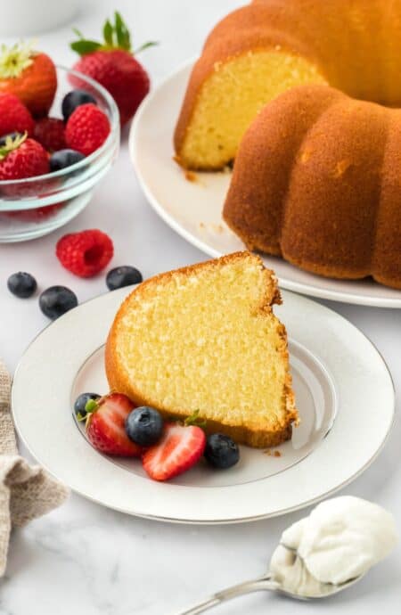 A slice of whipping cream pound cake served on a plate, garnished with fresh strawberries and blueberries, with the rest of the cake and a bowl with more fruit in the background