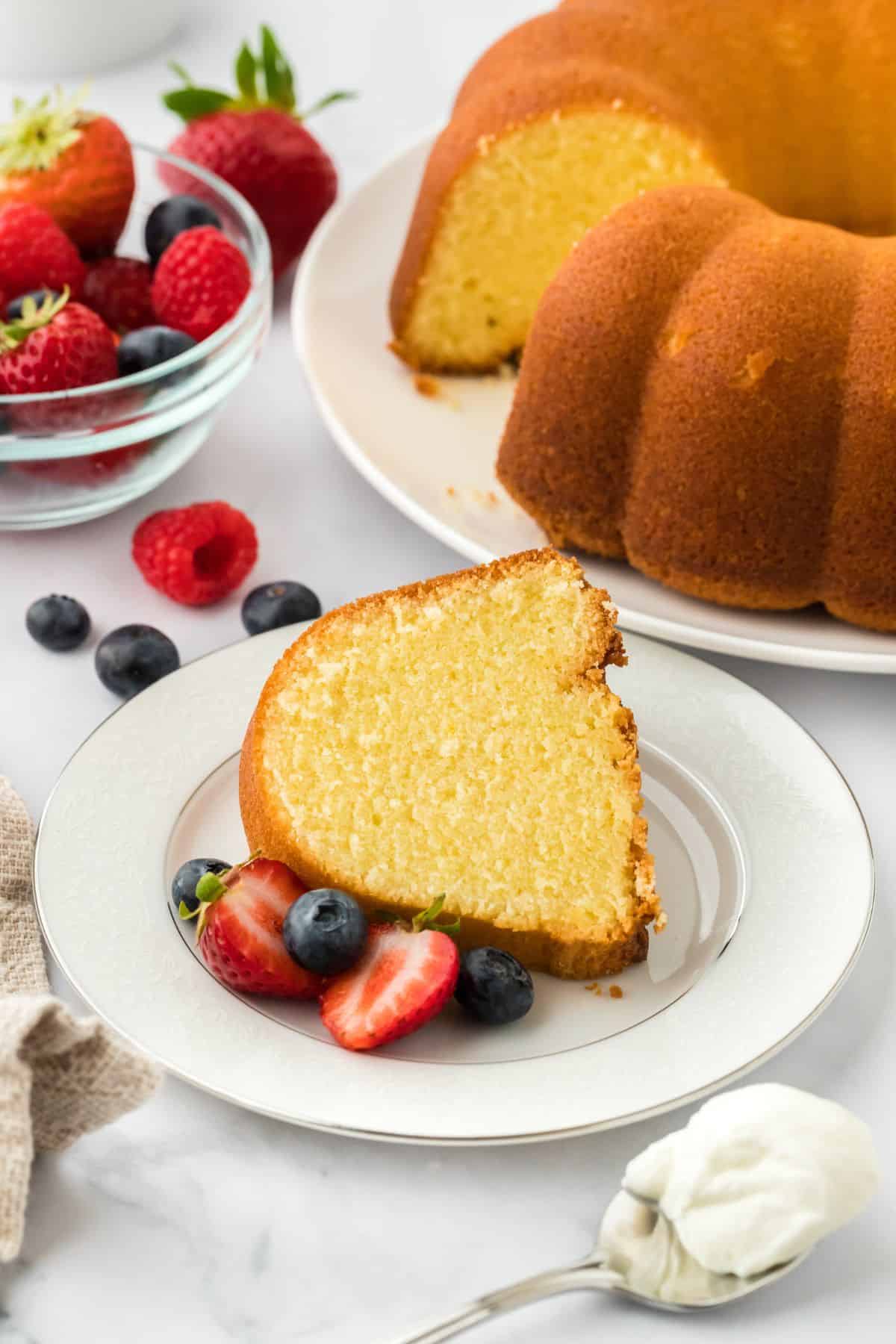 A slice of whipping cream pound cake served on a plate, garnished with fresh strawberries and blueberries, with the rest of the cake and a bowl with more fruit in the background