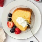 A plated slice of whipping cream pound cake topped with whipped cream and berries, with scattered berries around the plate and a bowl with whipped cream in the background