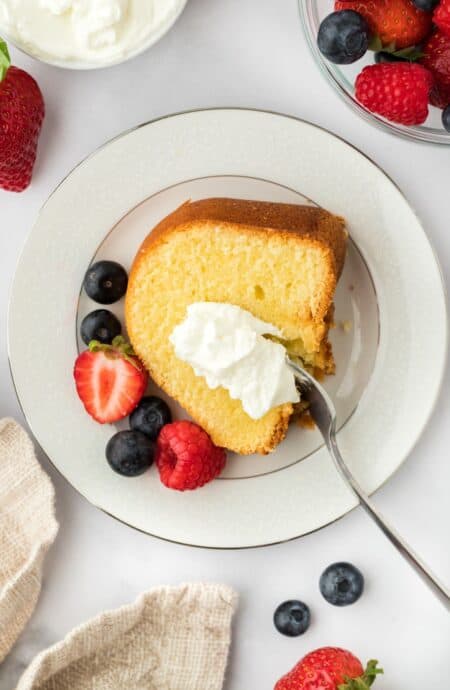 A plated slice of whipping cream pound cake topped with whipped cream and berries, with scattered berries around the plate and a bowl with whipped cream in the background