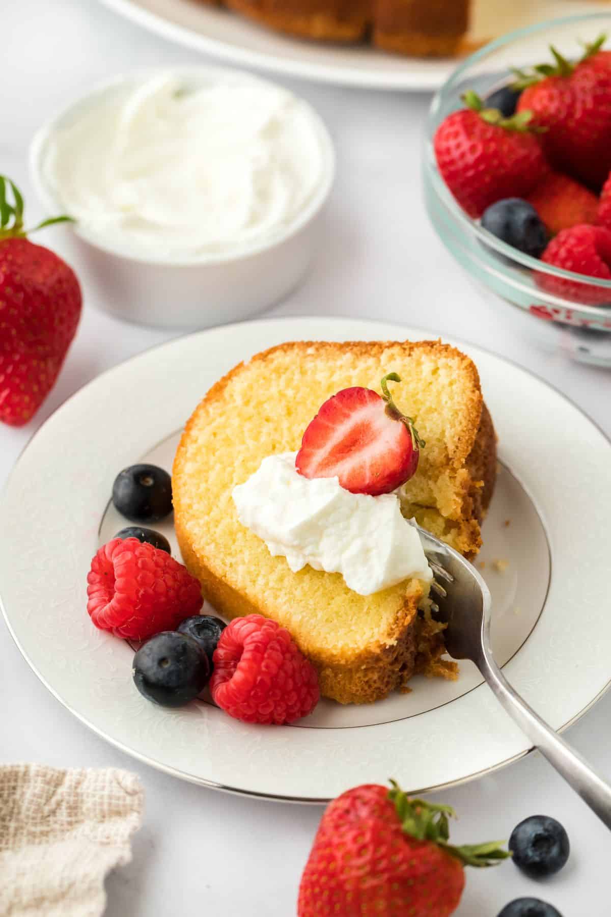 A plated slice of whipping cream pound cake topped with whipped cream and fresh strawberries, with scattered berries around the plate and a bowl with whipped cream in the background