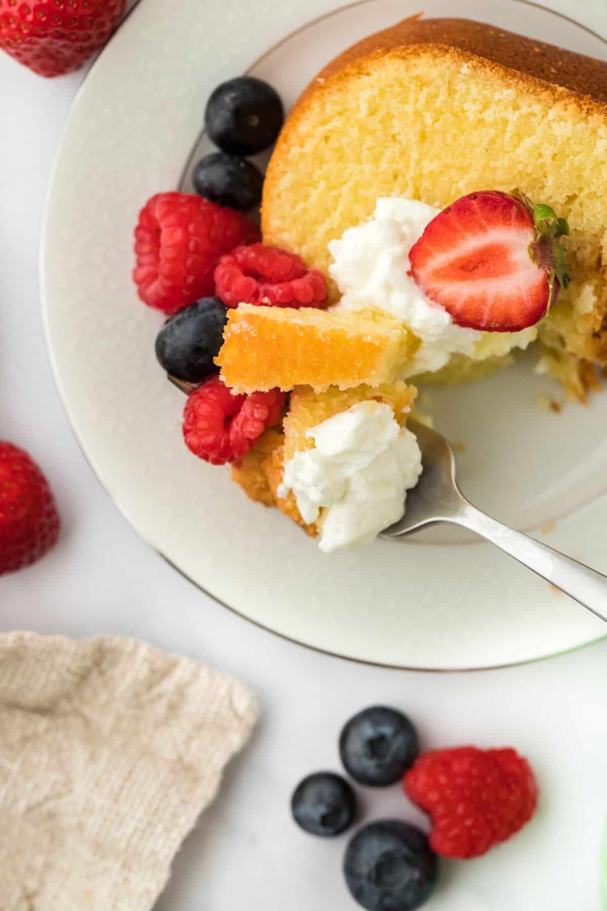 Overhead shot of a fork holding a bite of whipping cream pound cake topped with whipped cream and fresh fruit, placed on a plate with berries in the foreground