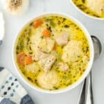 Overhead view of two bowls of chicken and dumpling soup with carrots, celery, and dumplings, accompanied by fresh herbs and garlic as garnish