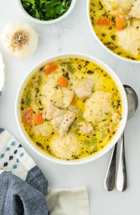 Overhead view of two bowls of chicken and dumpling soup with carrots, celery, and dumplings, accompanied by fresh herbs and garlic as garnish