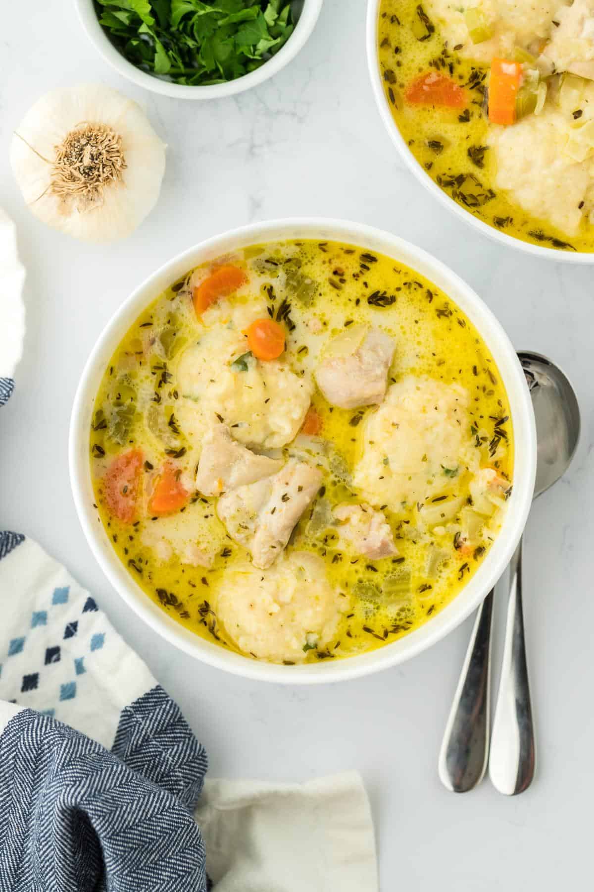 Overhead view of two bowls of chicken and dumpling soup with carrots, celery, and dumplings, accompanied by fresh herbs and garlic as garnish