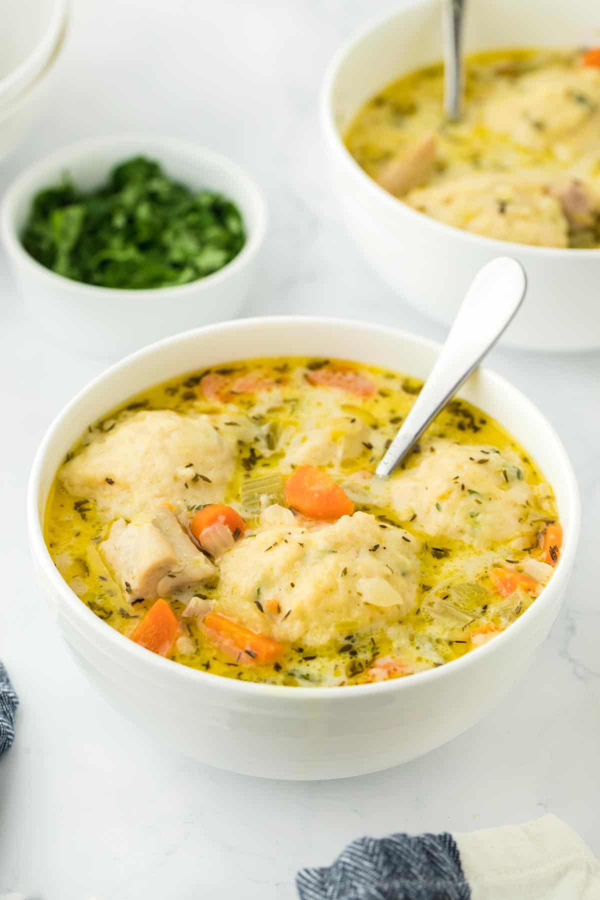 A side view of two bowls of chicken and dumpling soup on a white table, with spoons and a garnish bowl nearby