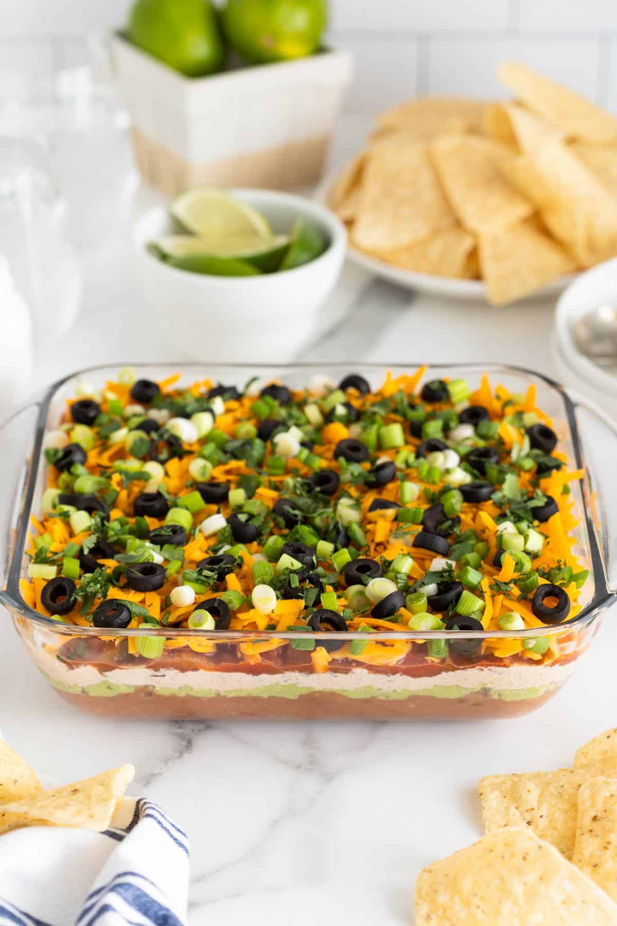 7 layer bean dip with chips and limes in the background on white countertop