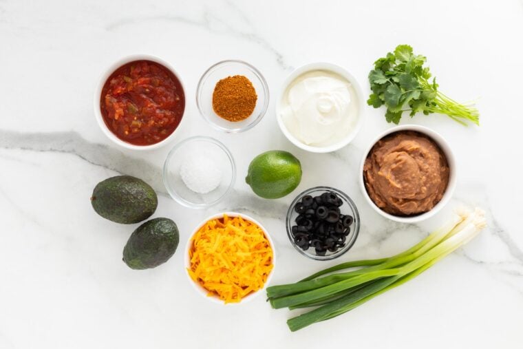 Refried beans, cheeses, salsa, sour cream, in glass bowls on white countertop