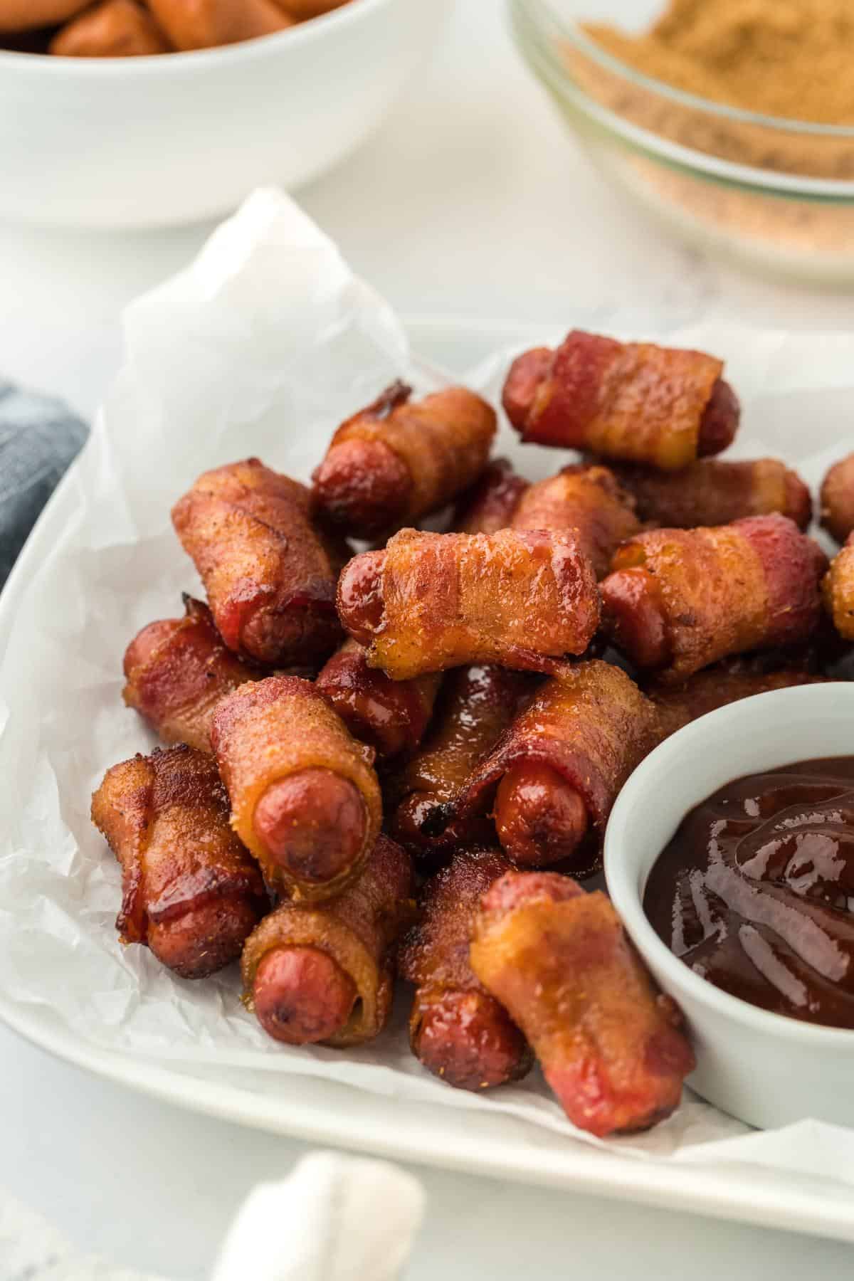 A closeup of a pile of bacon wrapped smokies served with a side of barbecue sauce in a small bowl