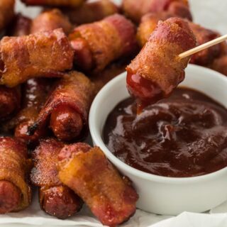 Bacon wrapped smokies on a plate with a smokie being dipped in barbecue sauce with a toothpick