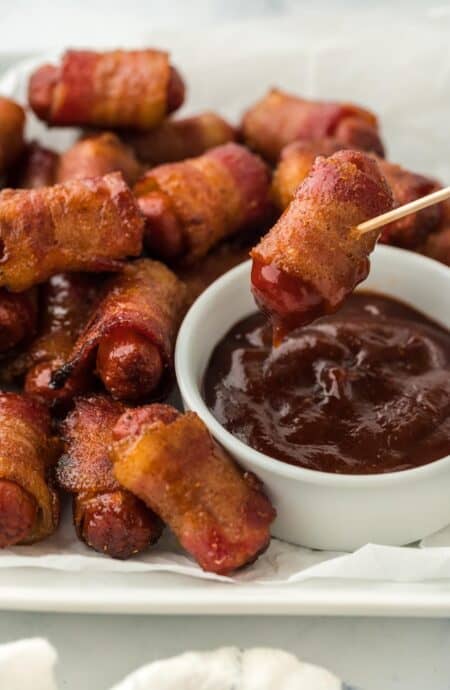 Bacon wrapped smokies on a plate with a smokie being dipped in barbecue sauce with a toothpick