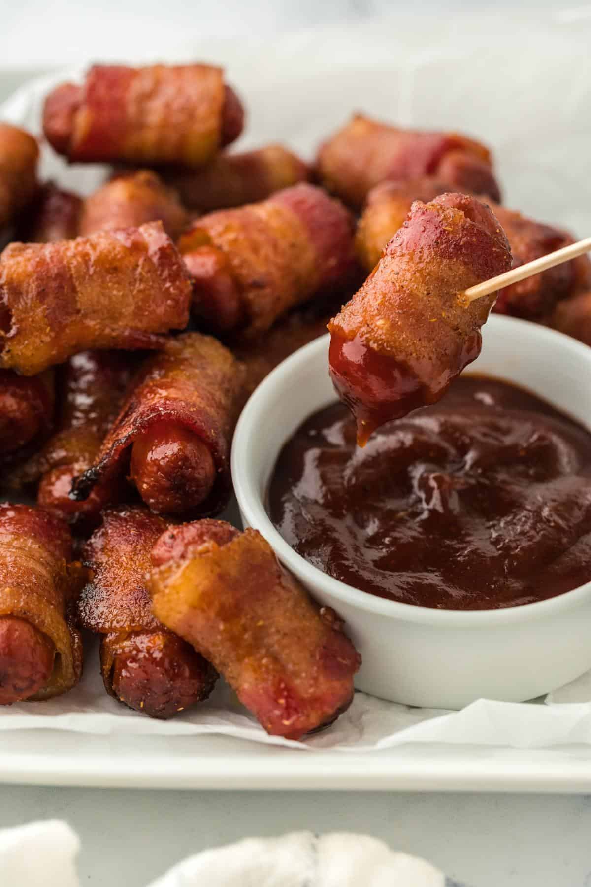 Bacon wrapped smokies on a plate with a smokie being dipped in barbecue sauce with a toothpick