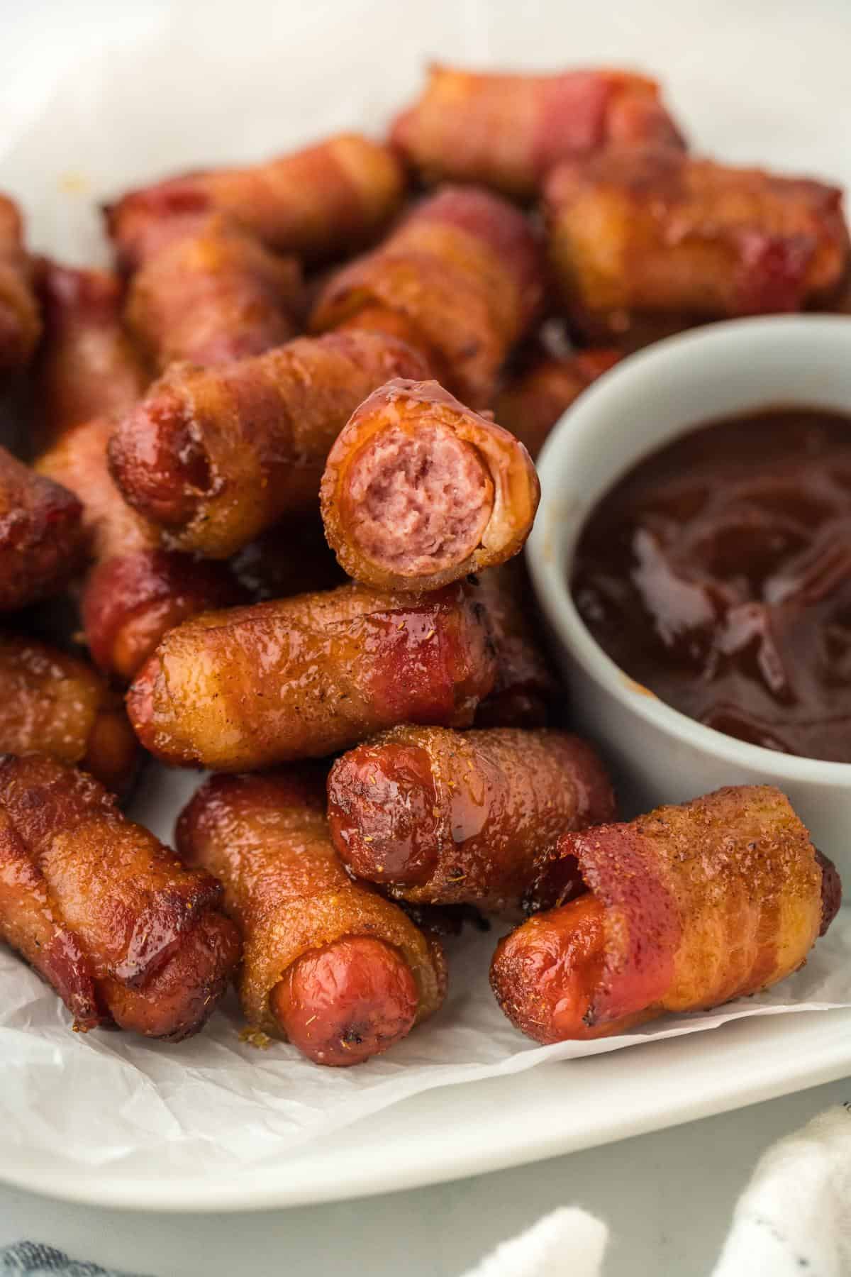 Closeup of bacon wrapped smokies, one with a bite taken out, showing the sausage filling next to a bowl of barbecue sauce