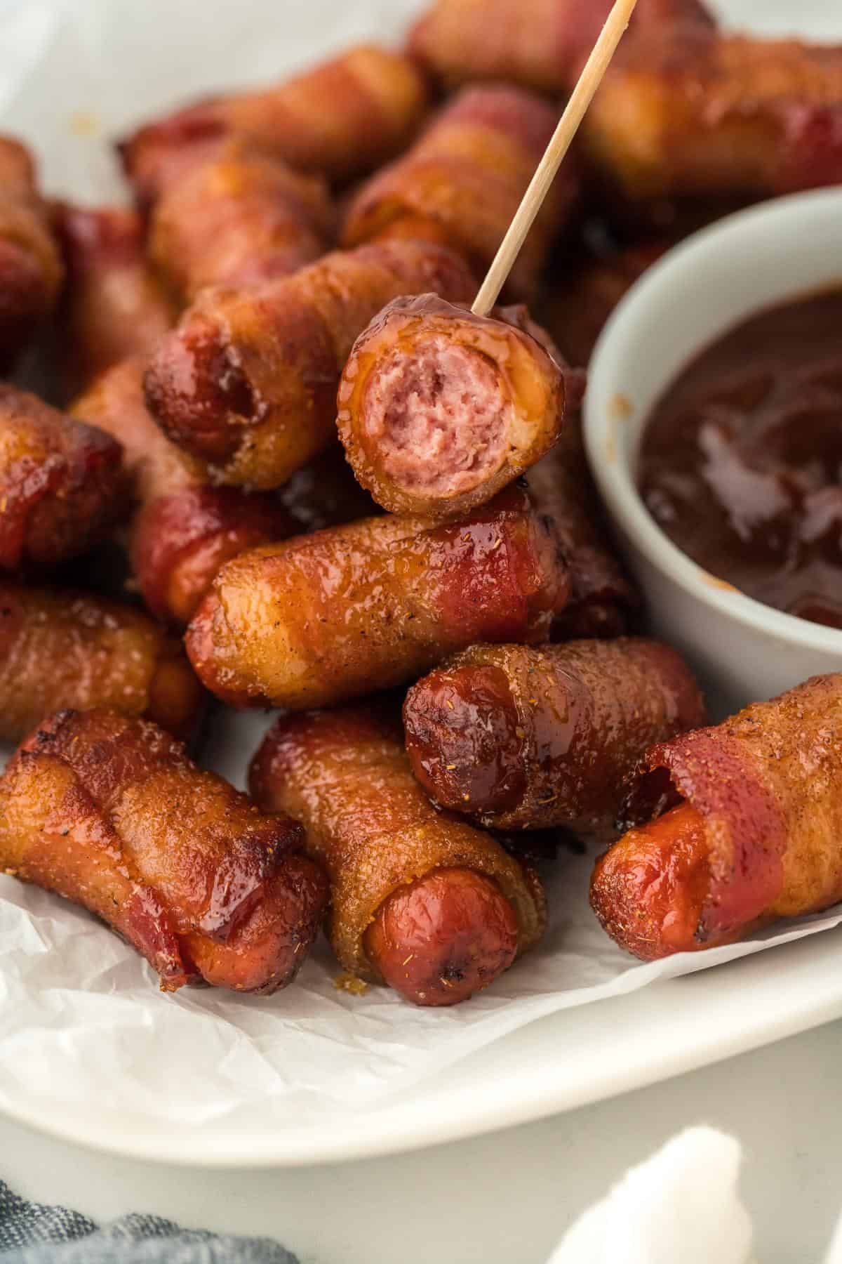 A bacon-wrapped smoky sausage on a toothpick being dipped in barbecue sauce