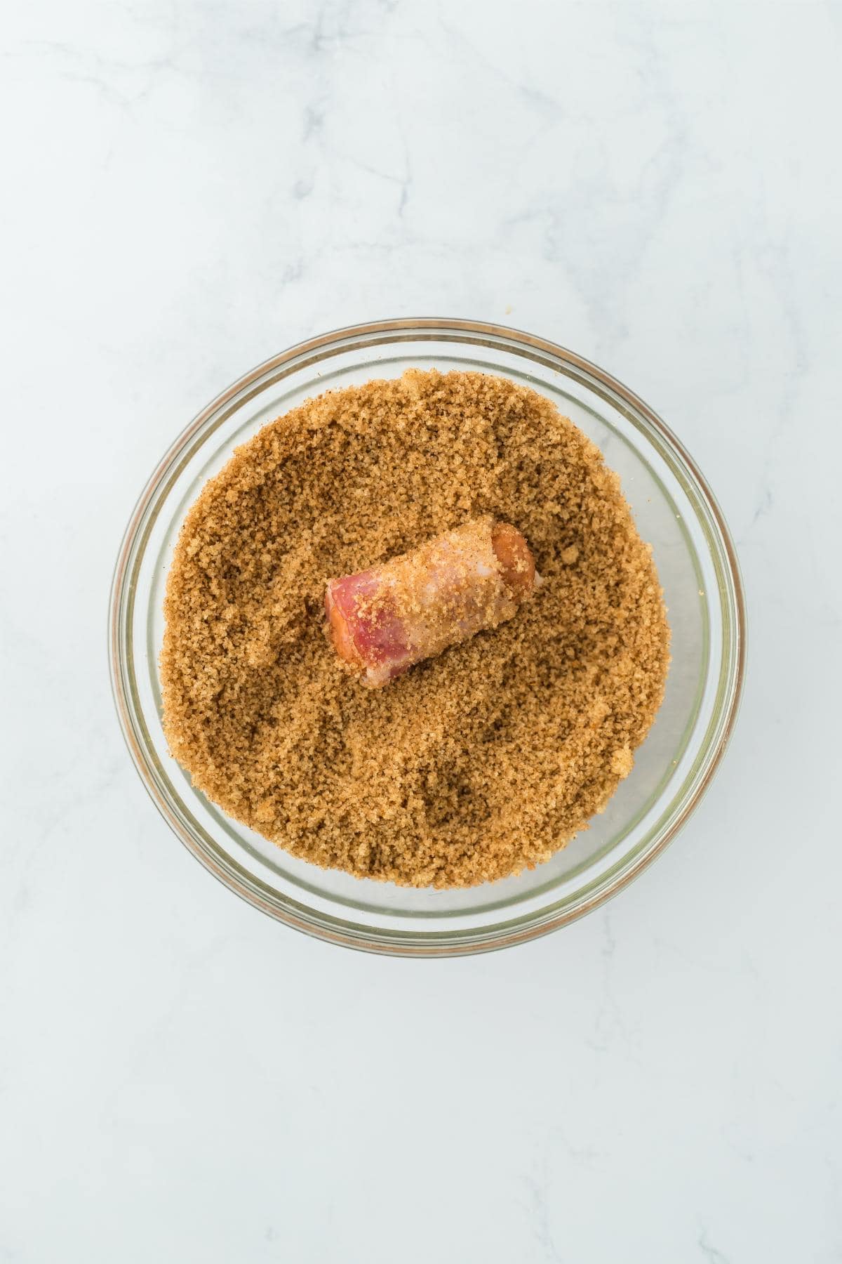 A bowl filled with a mixture of brown sugar and a bacon-wrapped sausage being rolled in the sugar