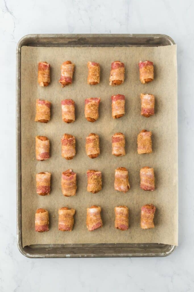 A baking sheet lined with parchment paper containing multiple bacon-wrapped sausages covered in brown sugar