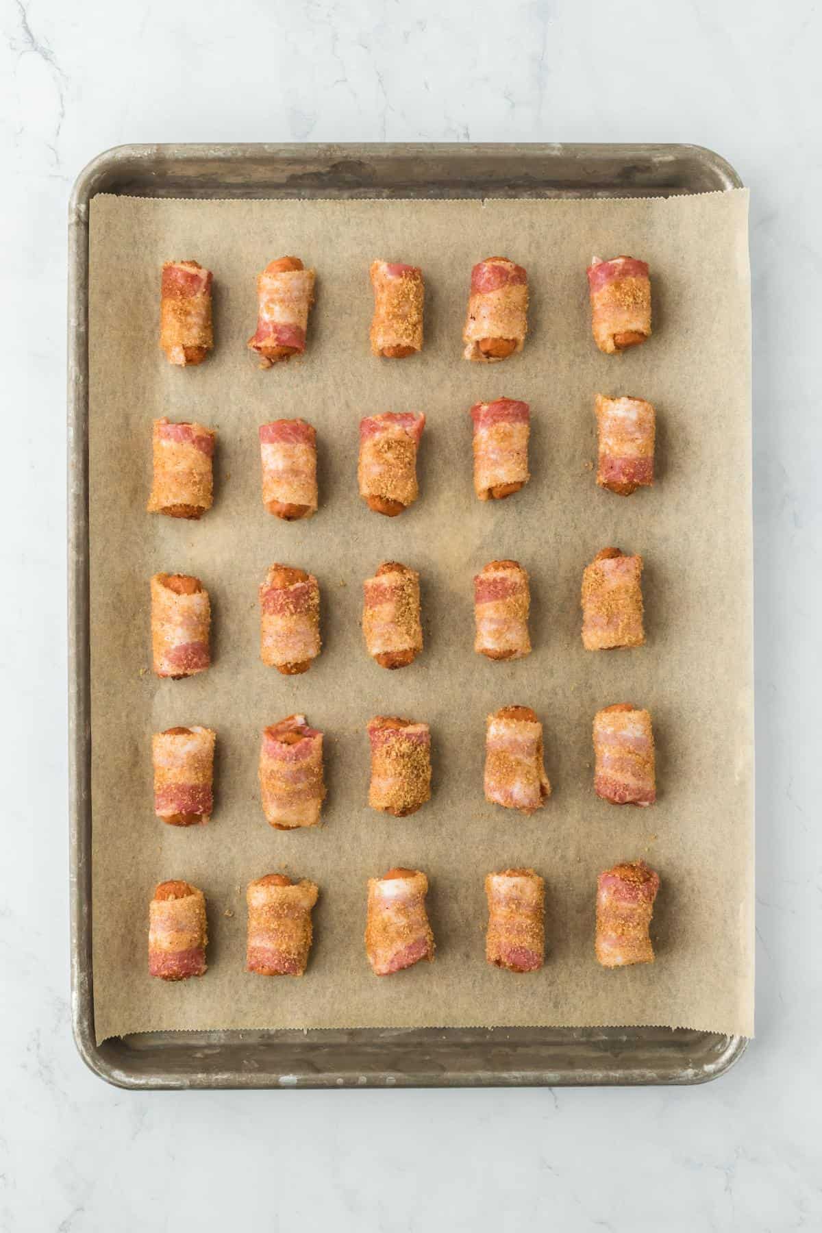 A baking sheet lined with parchment paper containing multiple bacon-wrapped sausages covered in brown sugar