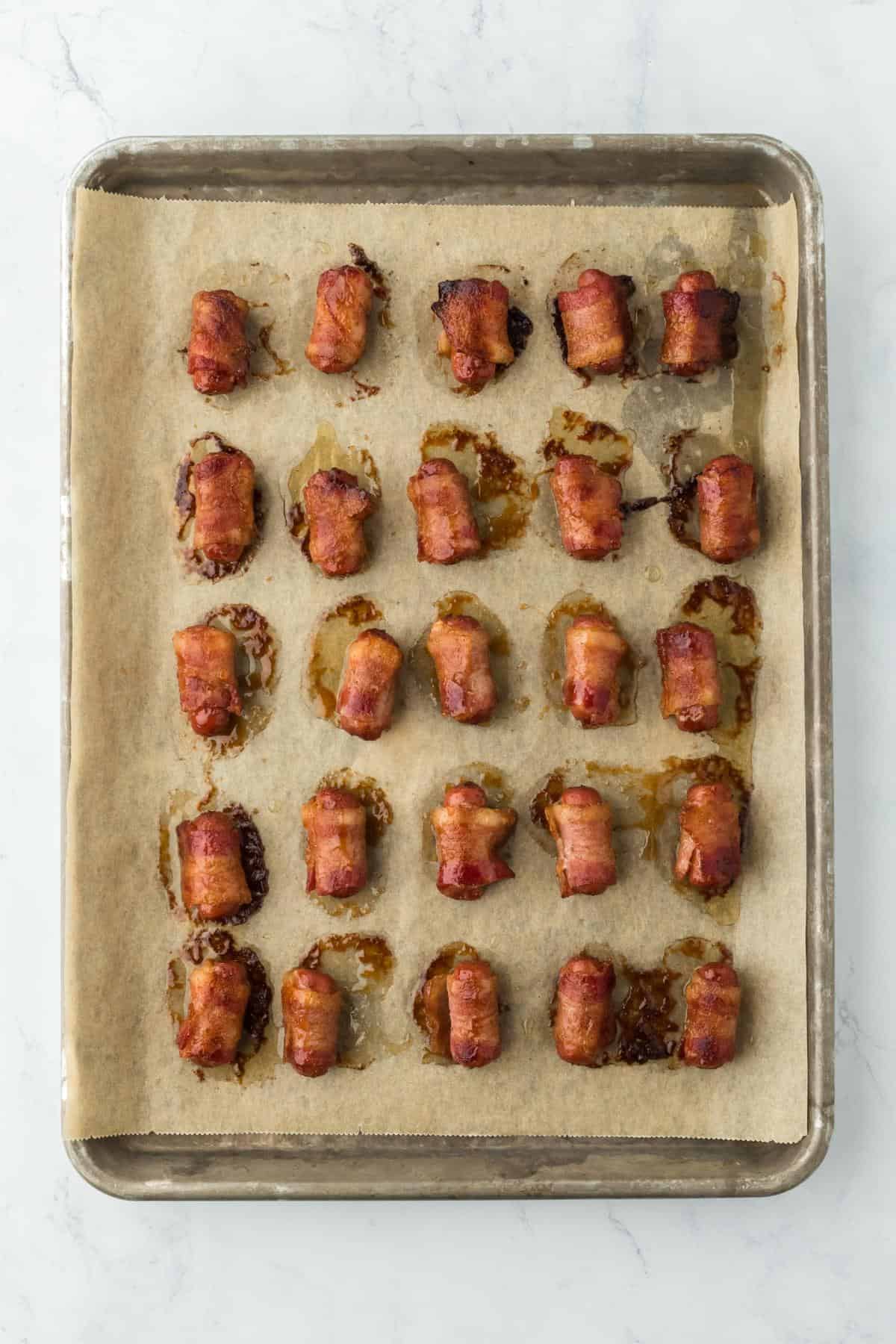 A closeup of baked bacon-wrapped sausages with some caramelized brown sugar and bacon grease on the baking sheet