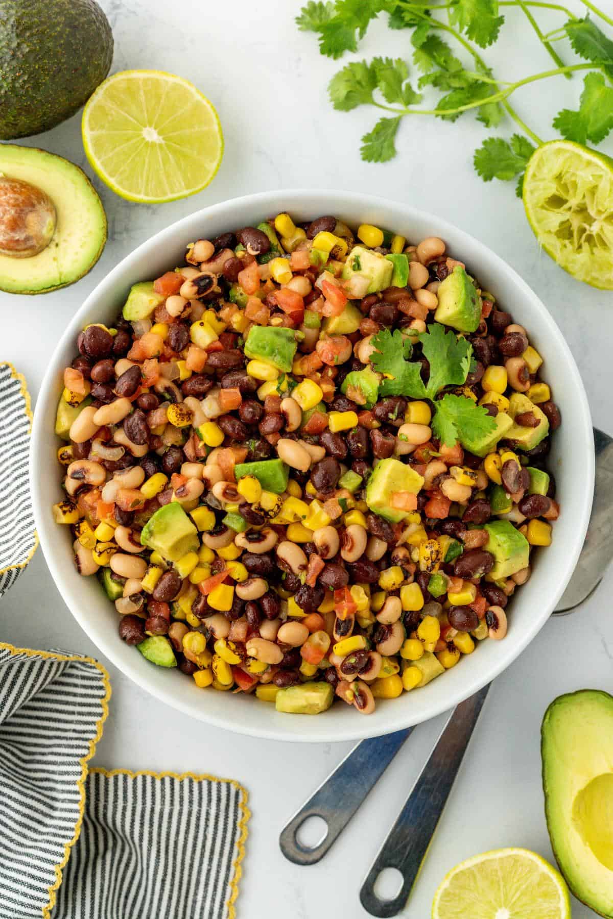 Overhead shot of cowboy caviar in a white bowl with lime wedges, cilantro, and avocado slices on the side